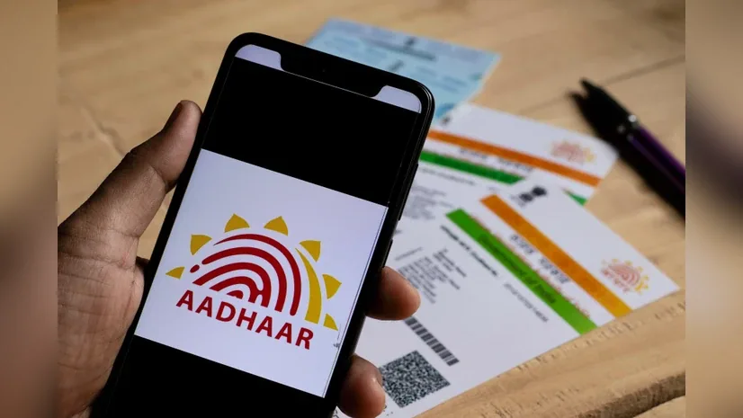 Hand holding smartphone displaying Aadhaar logo. Background shows Aadhaar cards and a pen on a wooden surface.