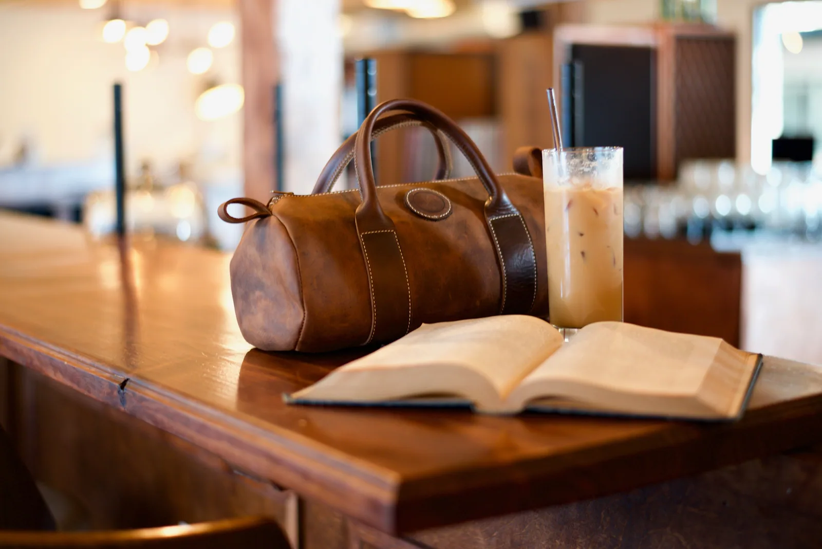 Brown leather bag and open book on a wooden table with iced coffee. Cozy, warm setting with blurred lights in the background.